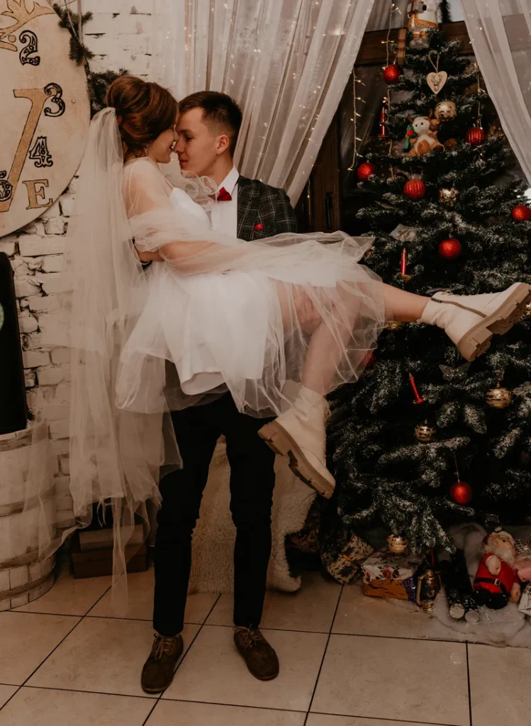A man carrying his bride in front of a beautifully decorated Christmas tree.