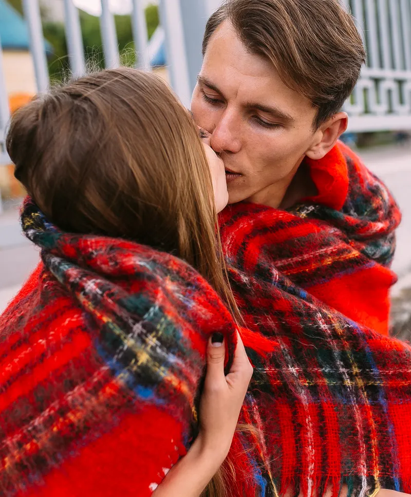 A picture of a couple kissing while wrapped in a warm plaid blanket.