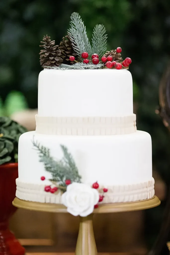 A white wedding cake with pinecone, evergreen and red berry decorations.