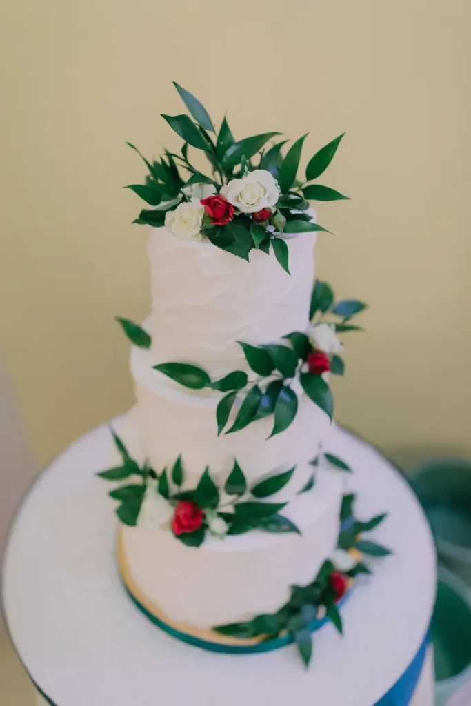 A textured white wedding cake with roses and prominent greenery. 