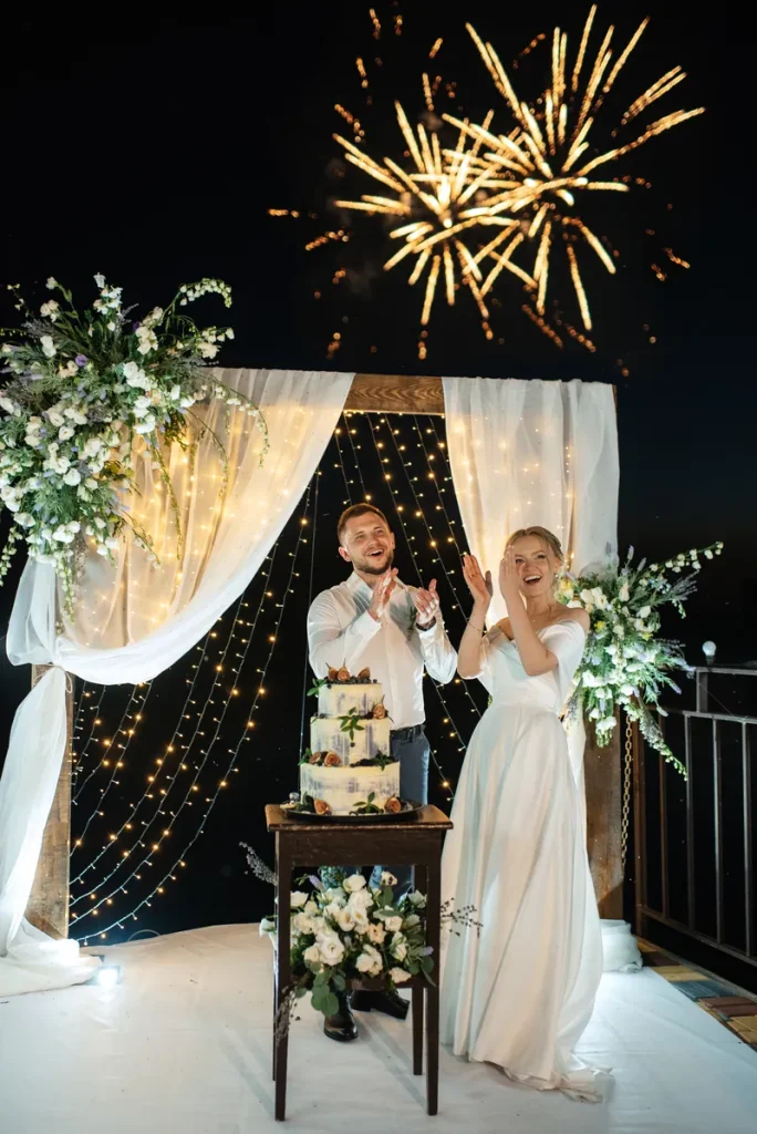 A happy couple in front of a wedding arch draped in white cloth and decorated with beautiful greenery and flowers.