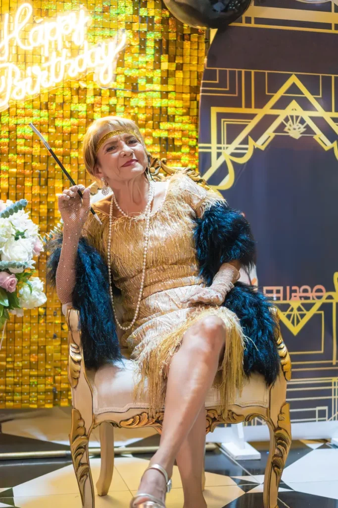 An older woman in front of an art deco background dressed in 1920's flapper attire.
